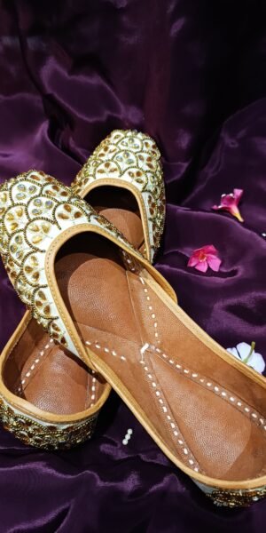 Ivory handcrafted Punjabi jutti with gold mirror work, zari embroidery, and a comfortable flat sole, ideal for bridal, festive, and traditional wear.