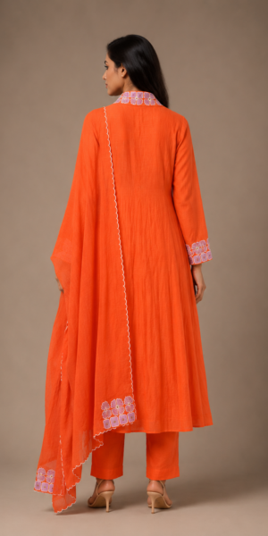 a saffron orange embroidered three-piece suit set featuring a gathered A-line kurta, straight cigarette pants, and a lightweight embroidered scalloped dupatta in a luxury studio setting.